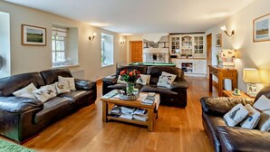 Ein Wohnzimmer mit zwei Ledersofas, einem Couchtisch mit Büchern, einem Kamin und einer sichtbaren Küche im Hintergrund.