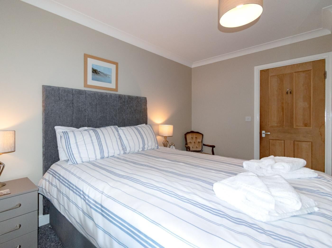 A neatly made bed with striped bedding, a bedside lamp, a chair, and a wooden door.