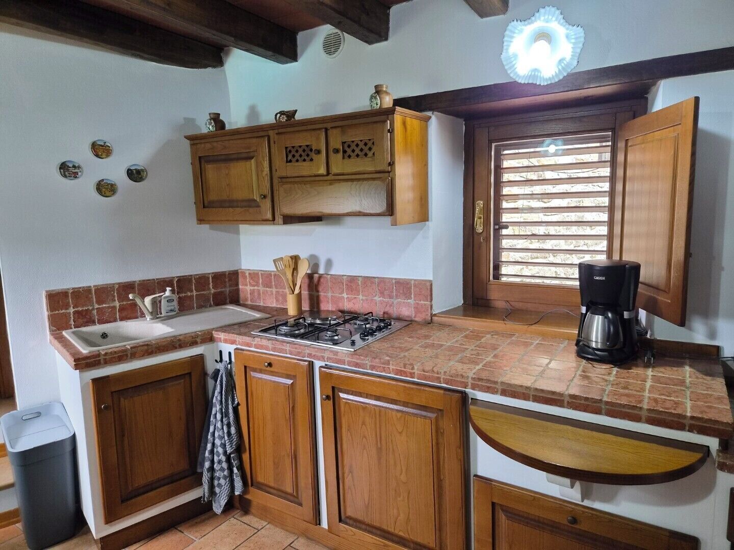Une cuisine avec des meubles de rangement en bois, une cuisinière à gaz, un évier, une machine à café et une fenêtre avec des stores.