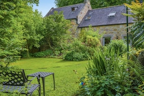 Ein Steinhaus mit gepflegtem Garten, Rasen und einem Metall-Tisch mit Stühlen.