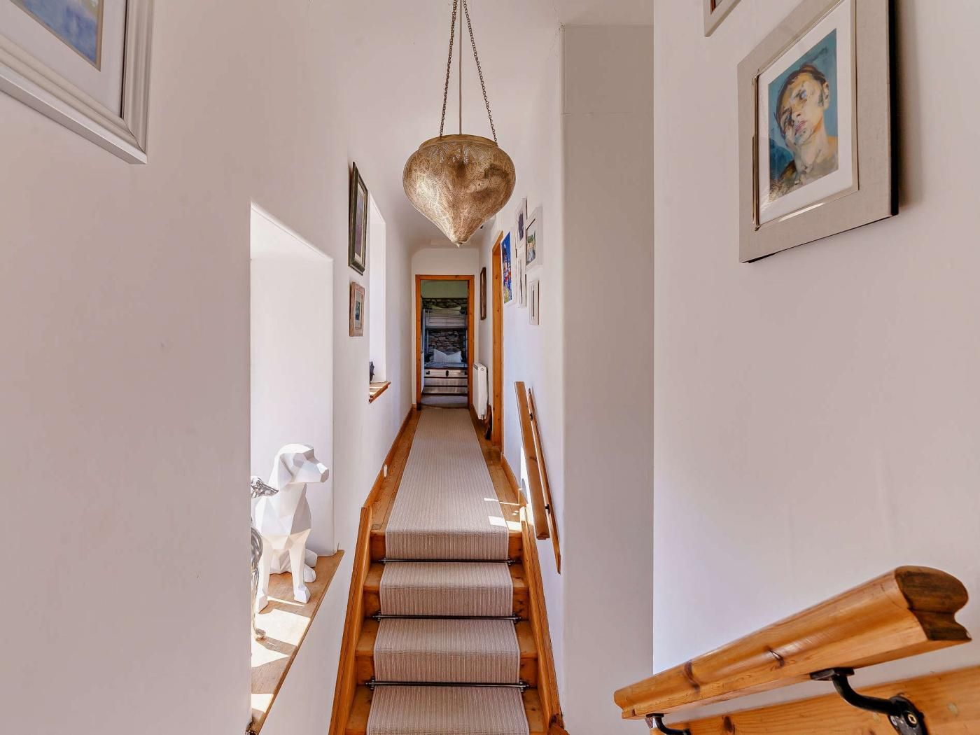 A hallway with a wooden staircase, a hanging light fixture, and framed artwork on the walls.
