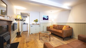 A living room with a sofa, a coffee table, and a fireplace. There is a kitchen in the background with a door leading to another room. - 2 Bed in Bideford (oc-59434) (Appledore)