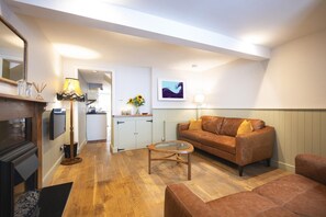 A living room with a sofa, a coffee table, and a fireplace. There is a kitchen in the background with a door leading to another room. - 2 Bed in Bideford (oc-59434) (Appledore)