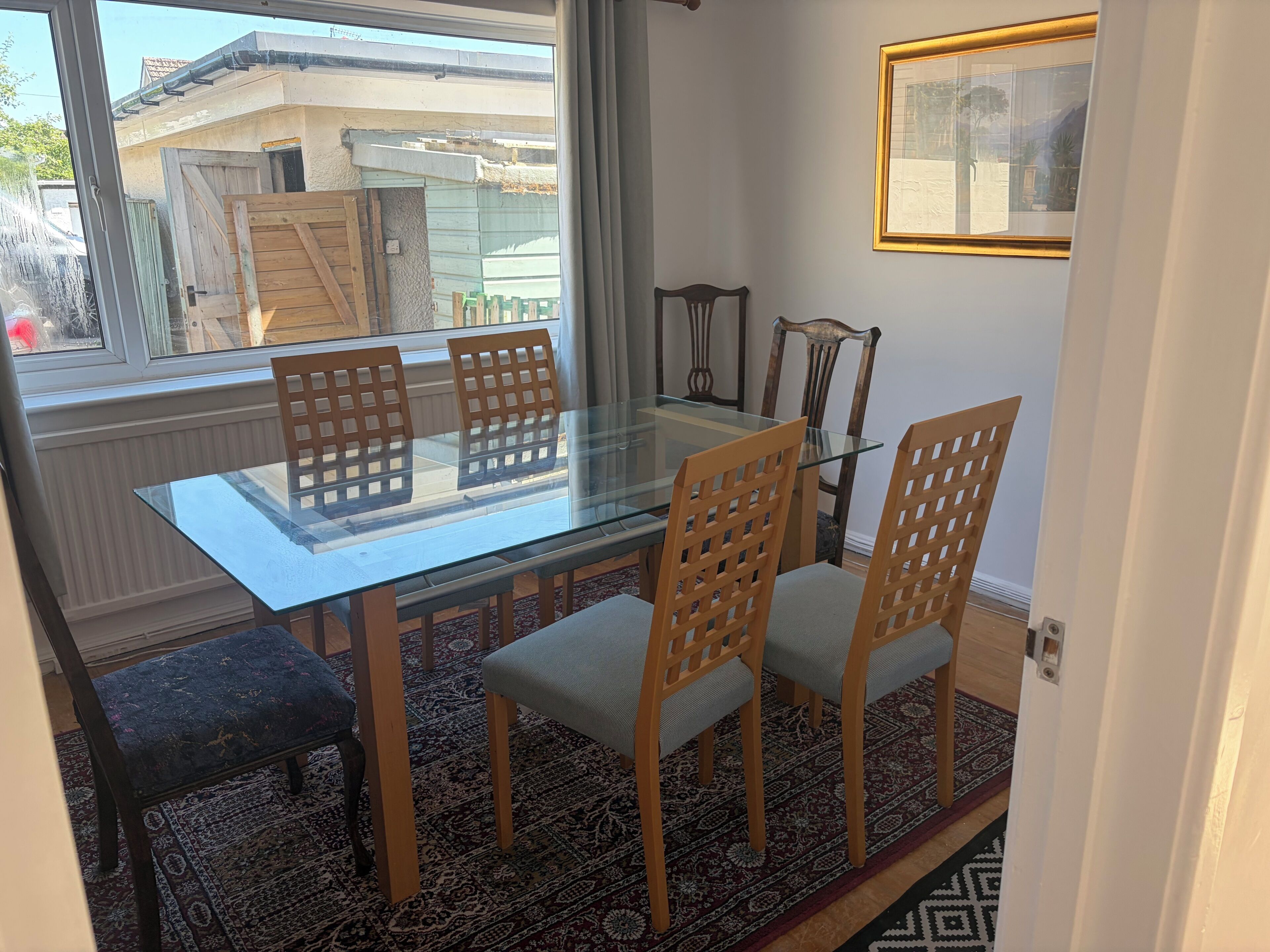 Een eetruimte met een glazen tafel, houten stoelen, een kamerbreed tapijt en uitzicht op buiten.