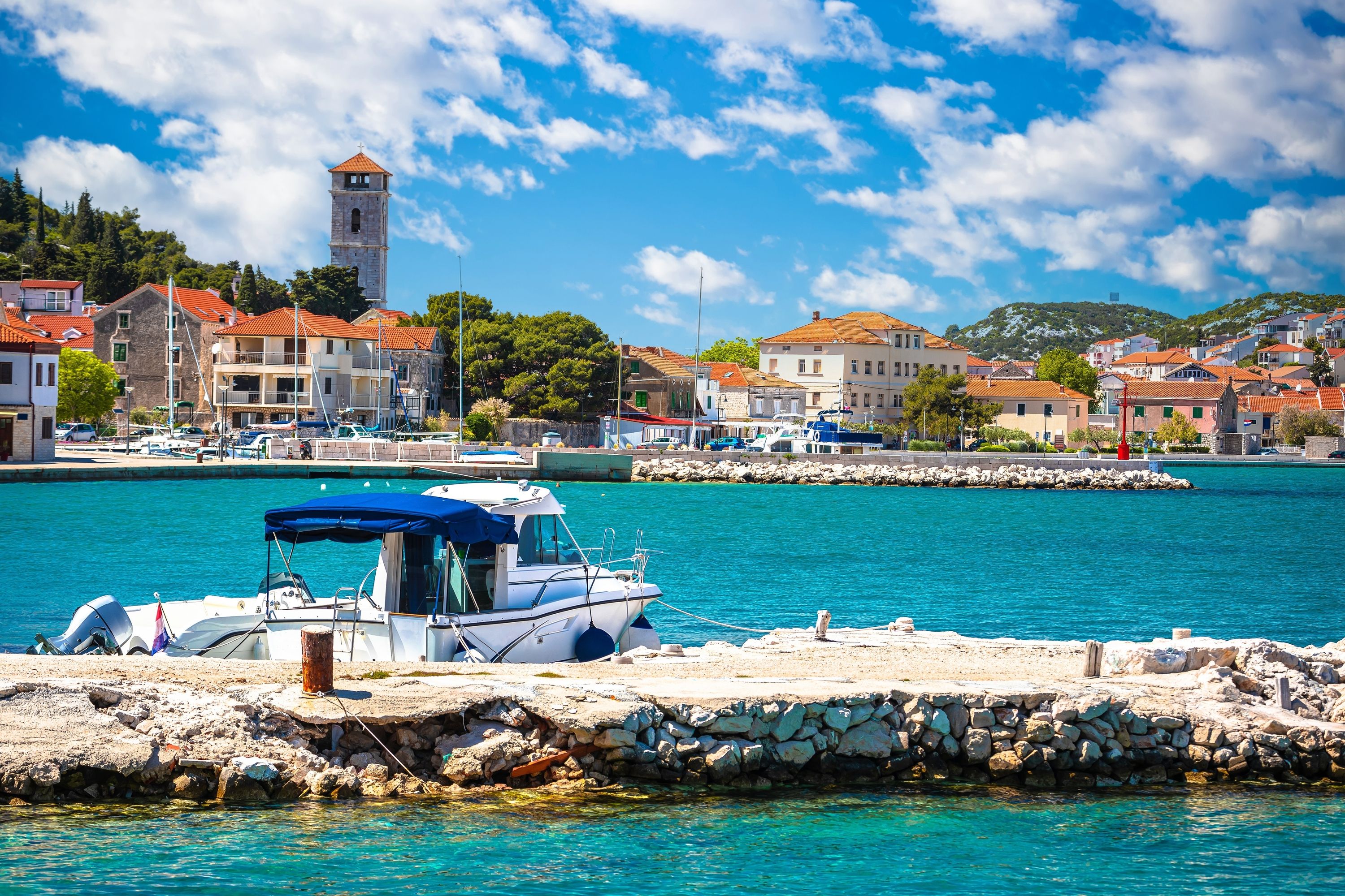 A coastal town with a clear blue sea, a stone pier, and buildings with red-tiled roofs.