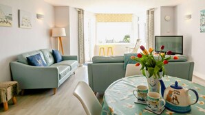 Ein modernes Wohnzimmer mit einer Couch, einem kleinen Tisch mit einer Tulpenvase, einem Fernseher und einem Fenster mit Vorhängen.
