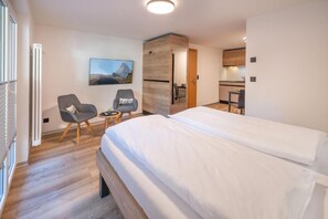 A modern hotel room with a large bed, two chairs, a small table, a painting, and a kitchen area. - Haus Juliana Alpenrose (Zermatt)