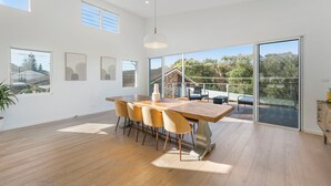 A spacious dining area with a large wooden table, multiple chairs, and a view of a house through large sliding glass doors.