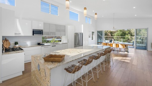 A modern kitchen with white cabinetry, a large island with bar stools, stainless steel appliances, and large windows.