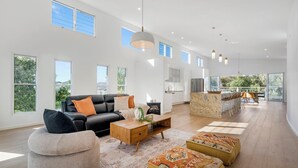 A spacious living area with a black leather sofa, wooden coffee table, and ottomans, large windows, and a kitchen area with a dining set.