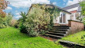 Ett bostadshus med en trappa av tegel som leder upp till en vit dörr, omgivet av en välskött gräsmatta och tät vegetation.