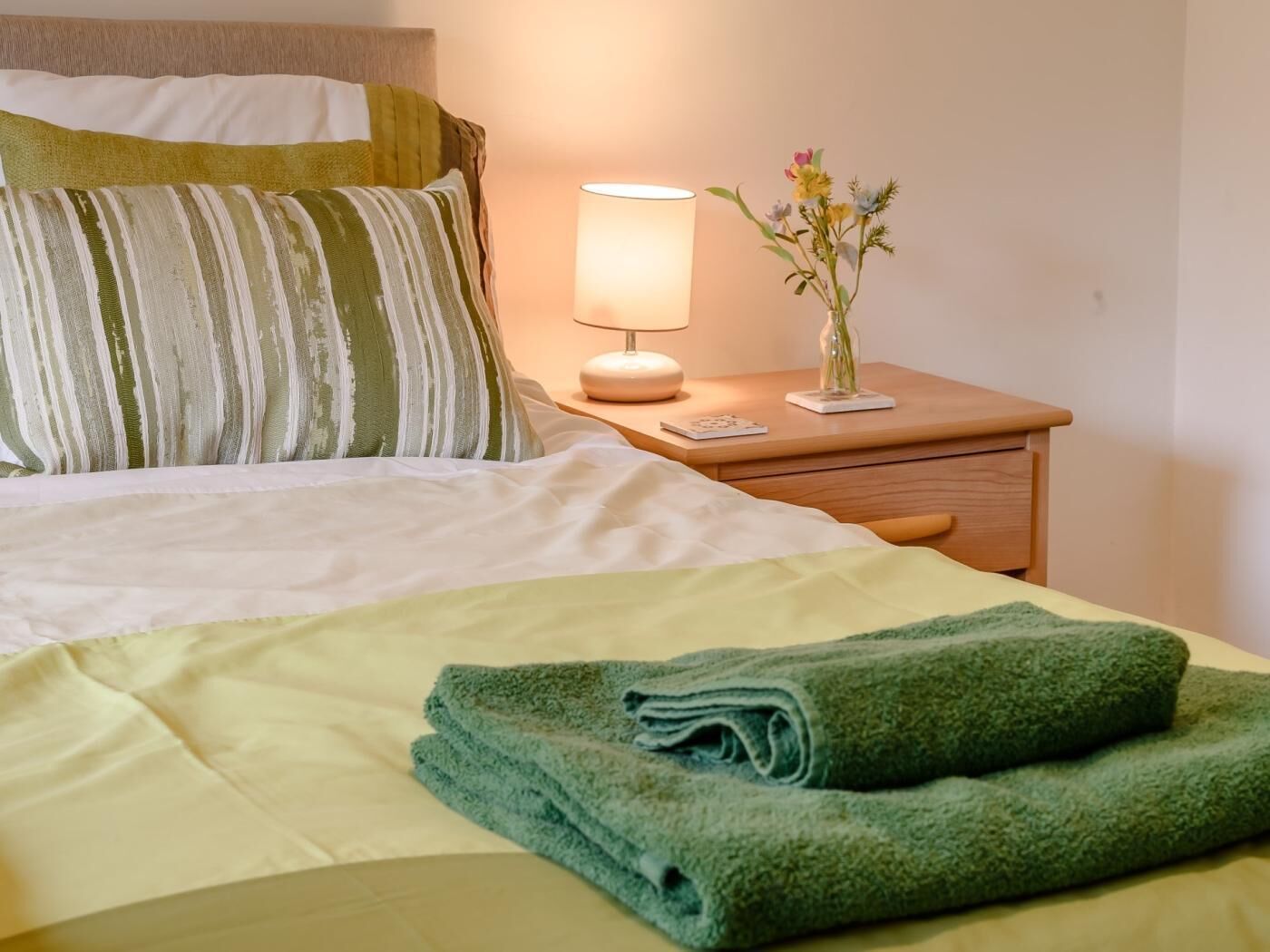A neatly made bed with green towels, a striped pillow, a bedside lamp, and a small vase with flowers on a wooden nightstand.