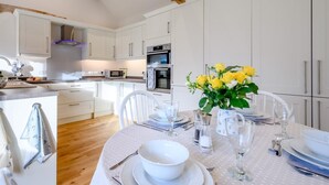 Een moderne keuken met witte kastjes, een houten balk in het plafond en een eethoek voor vier personen, gedekt met een witte tafelkleed en gele bloemen.