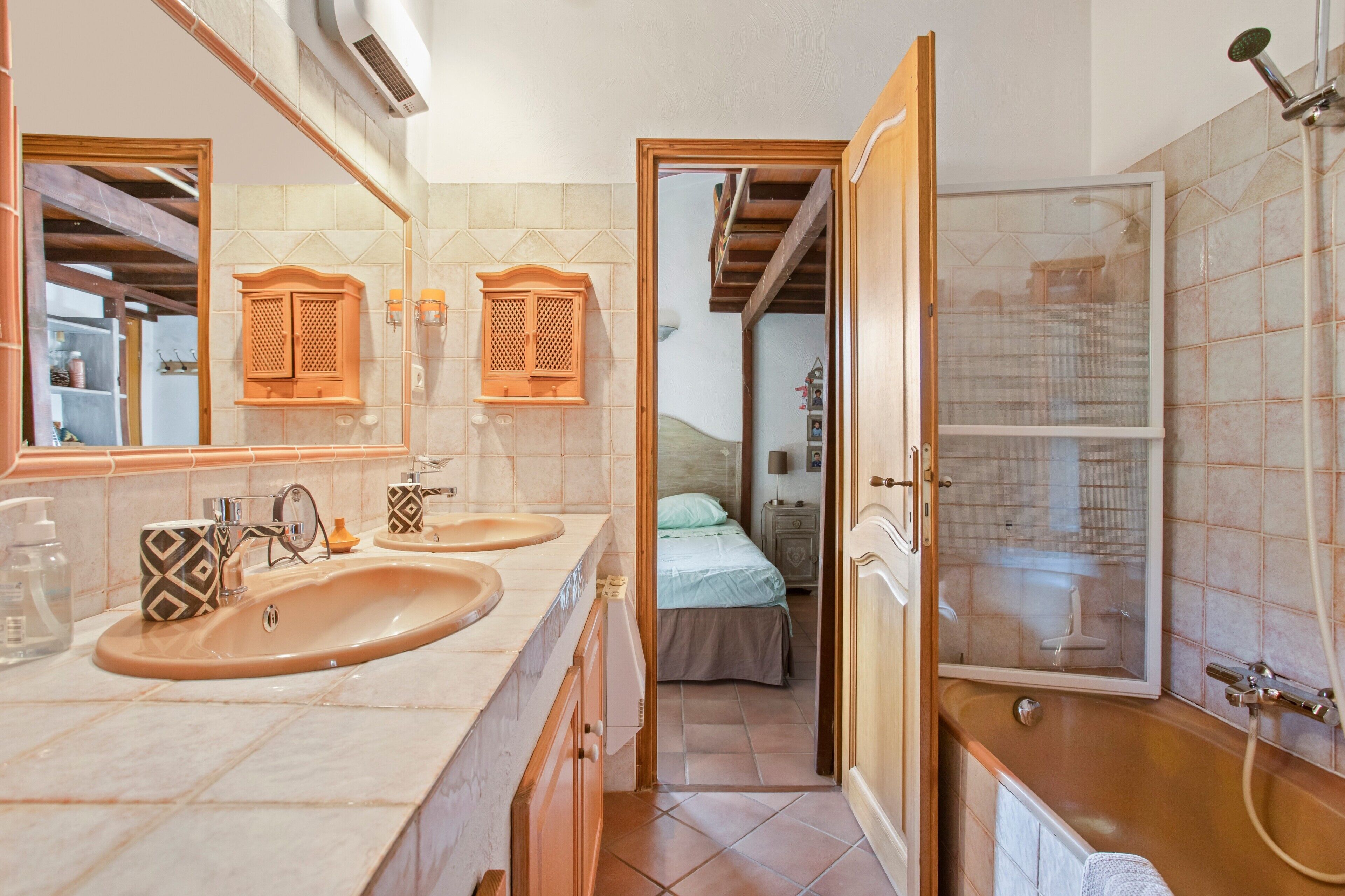 Ein Badezimmer mit zwei Waschbecken, einer Dusche und einem Bett, das durch eine geöffnete Tür sichtbar ist.