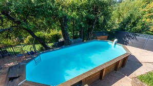 Una piscina rectangular amb un pont de fusta al voltant, situada en un jardí paisatjistic amb arbres i una tanca.