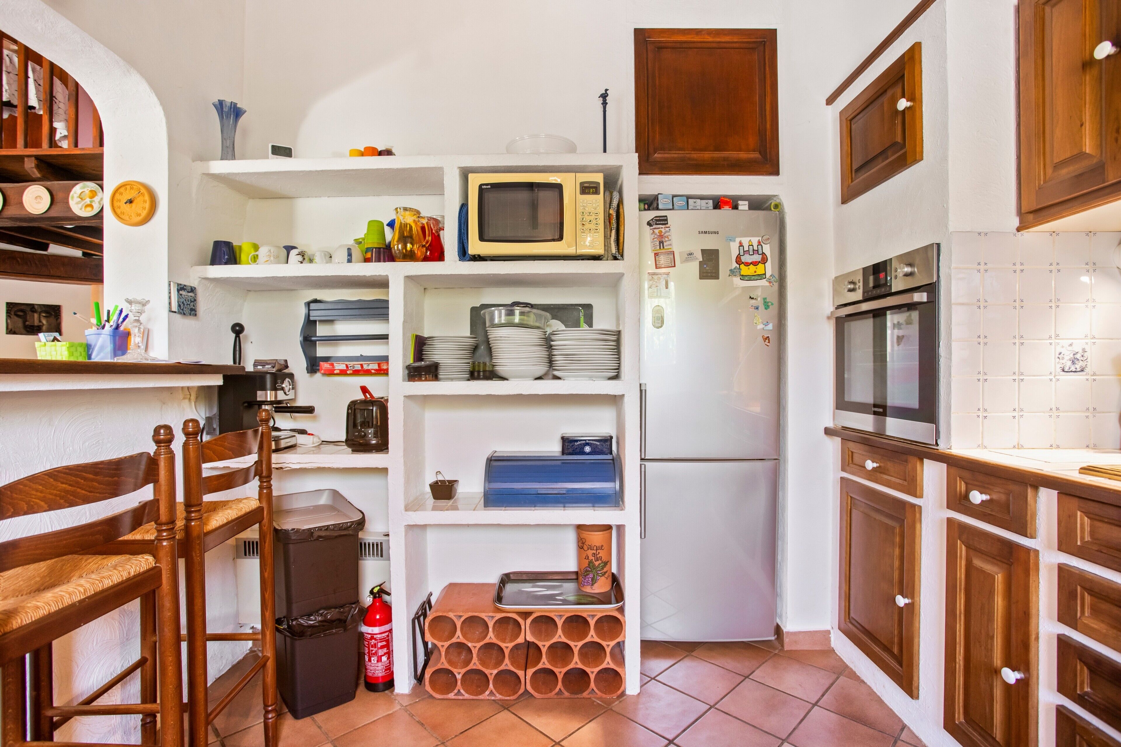 A compact kitchen with a microwave, refrigerator, and oven. There are shelves with dishes and a trash bin.