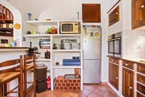 A compact kitchen with a microwave, refrigerator, and oven. There are shelves with dishes and a trash bin.