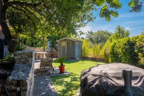 A backyard with a stone wall, a wooden bench, a small shed, and a covered grill area.
