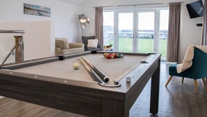 Ein Billardtisch mit Kugeln, eine Couch, ein Sessel, eine Lampe und ein Fernseher in einem Zimmer mit großen Fenstern.