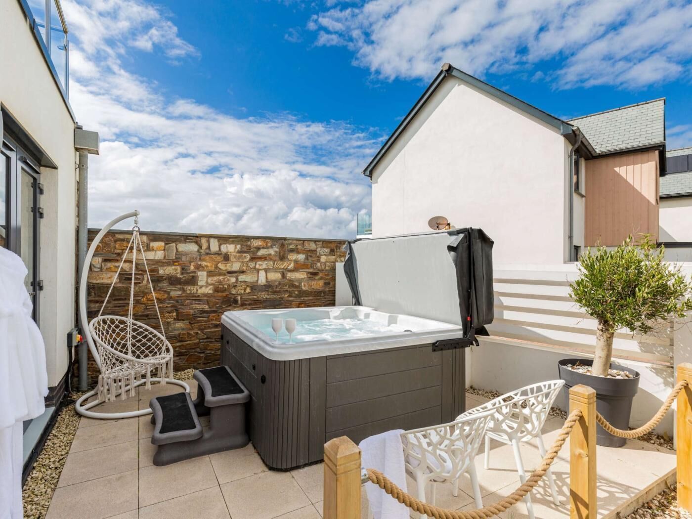 A hot tub in a patio area with a stone wall, a hanging chair, and a small tree.