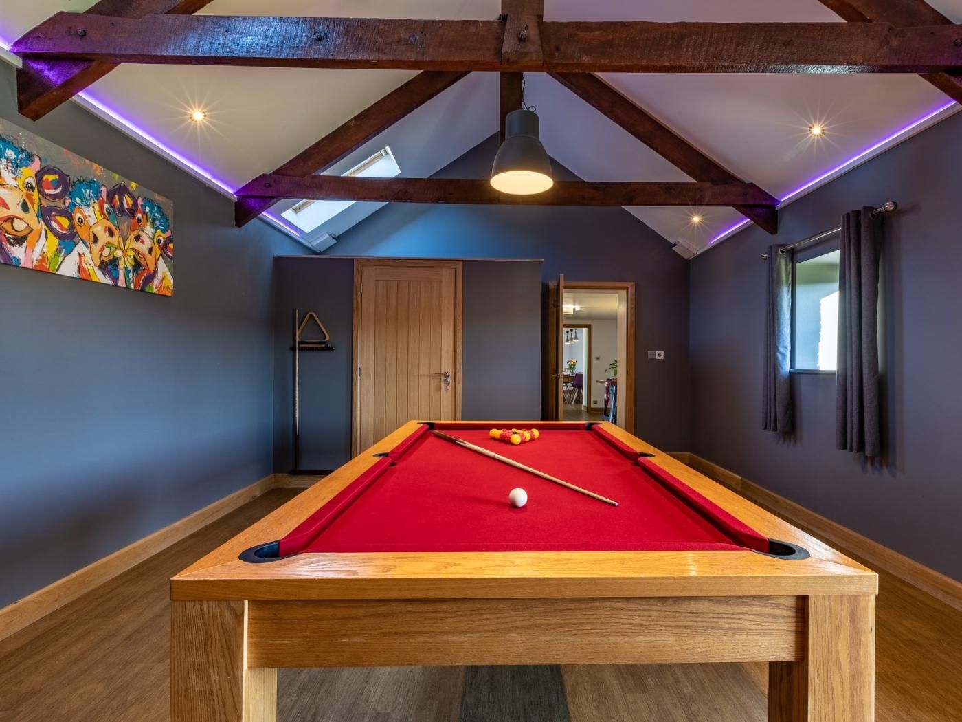 Une table de billard dans une pièce dotée d’un plafond en pente, de poutres en bois et d’un tableau coloré accroché au mur.