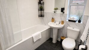 Un baño moderno con bañera, lavamanos e inodoro. Hay una repisa con artículos de aseo y una planta pequeña.