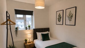 Un dormitorio con cama, mesita de noche, lámpara, planta colgante y ventana.