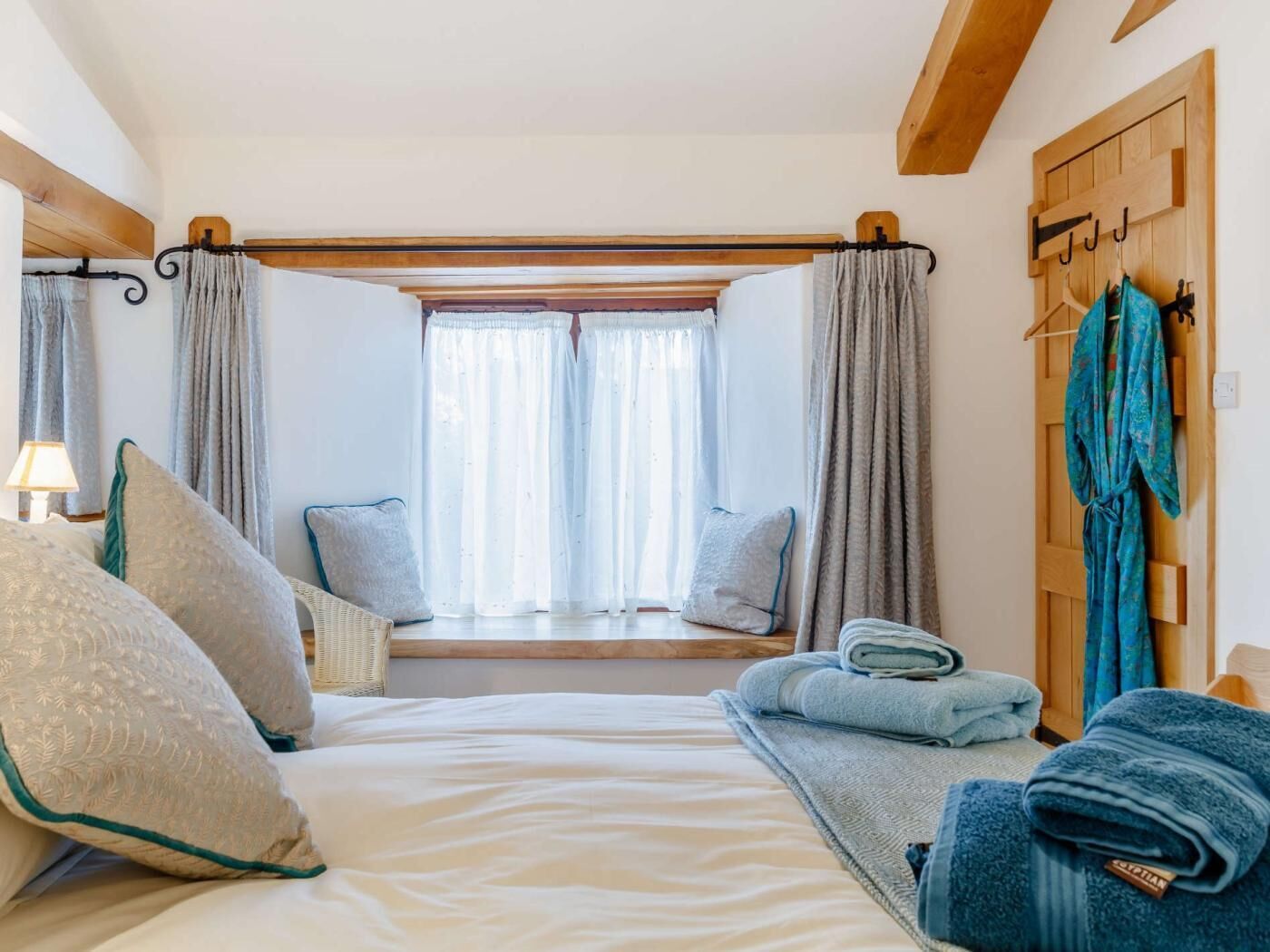 A bedroom with a bed, pillows, a window with curtains, a wooden headboard, and a towel rack.