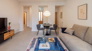 A modern living room with a sofa, a coffee table with a book and remote, a TV on a stand, and a dining area with chairs and a table. - Welcomer La Latina (Madrid)