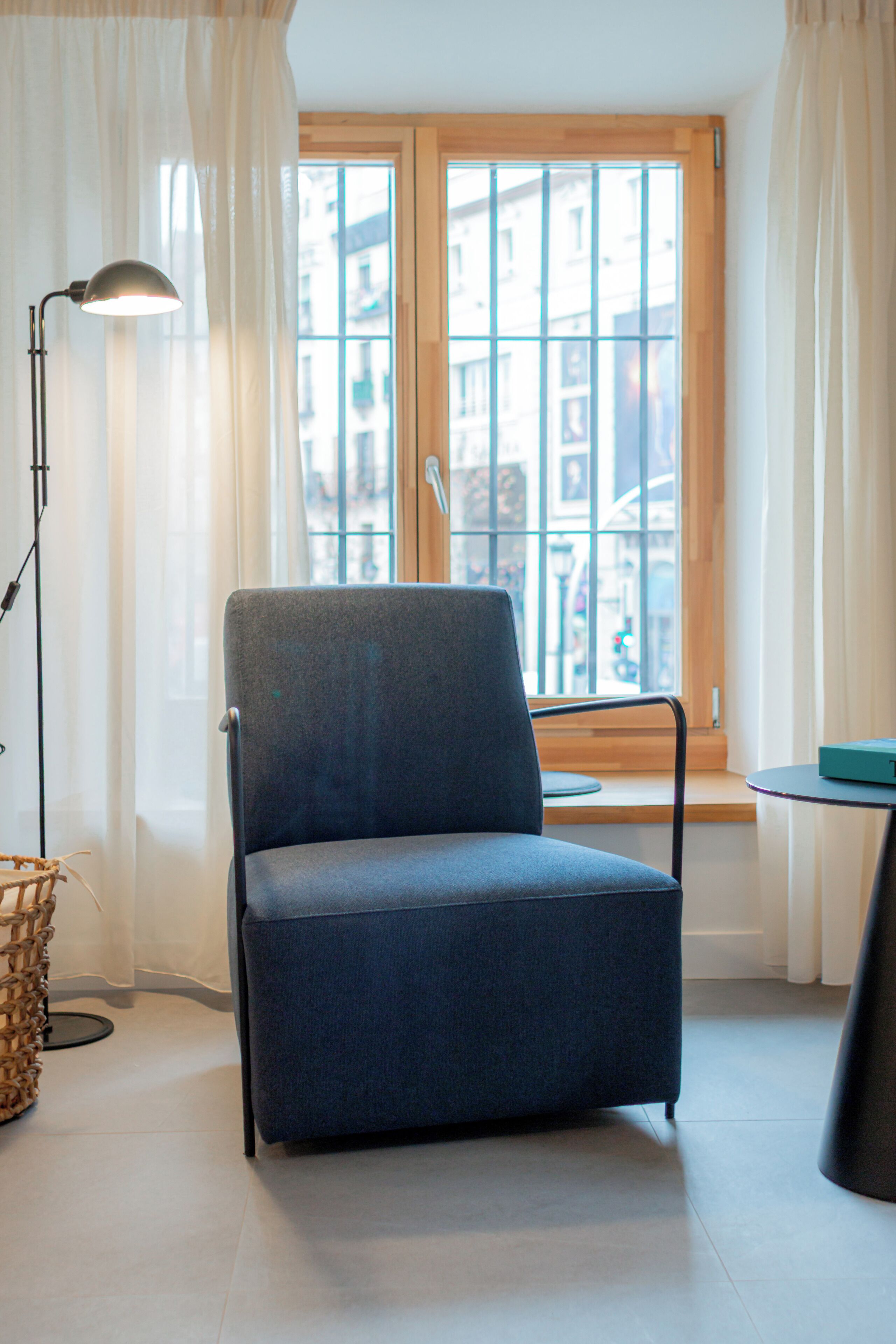 Een kamer met een blauwe fauteuil, een vloerlamp, een tafeltje en uitzicht door een raam met gordijnen.