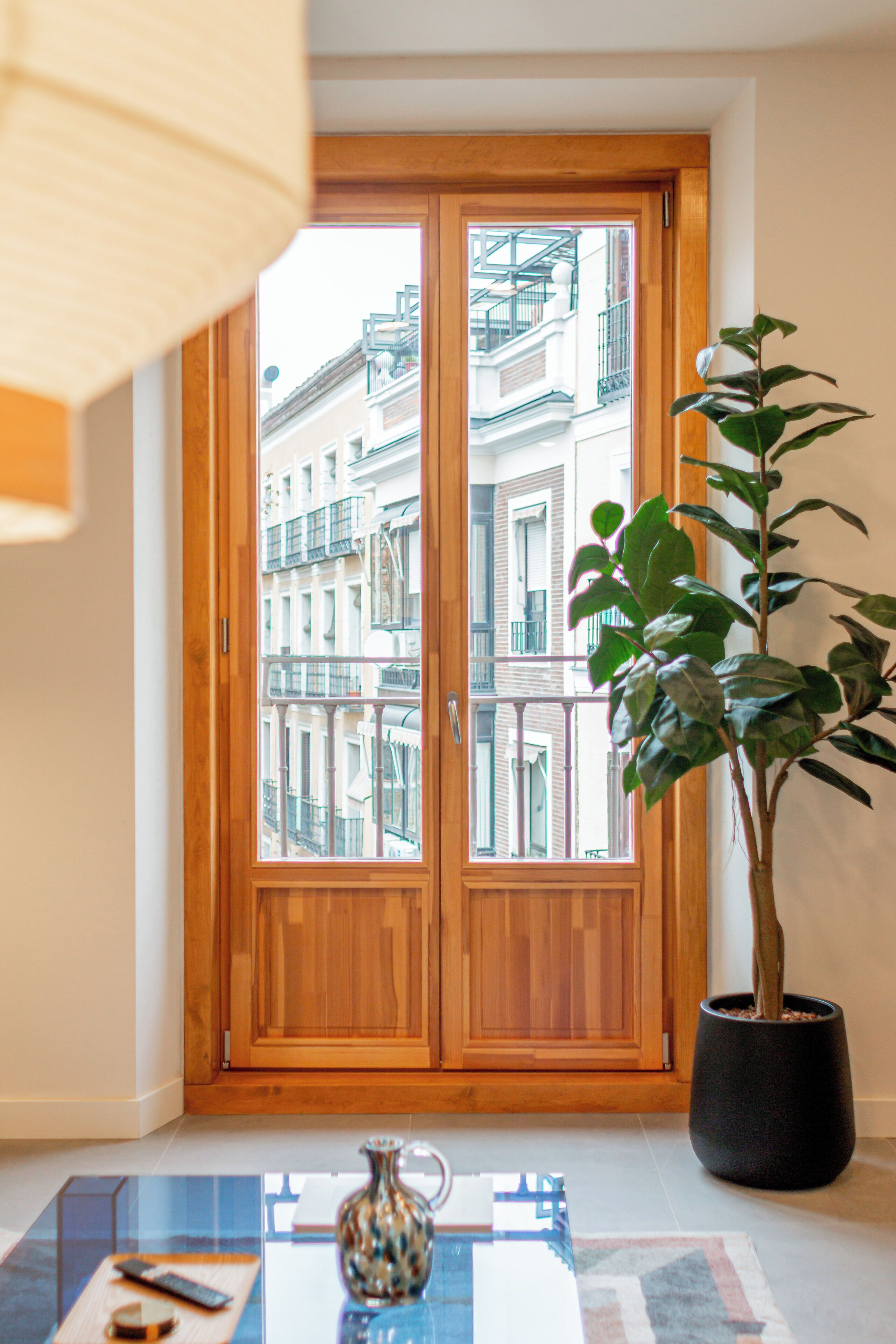 Una habitación con puerta de madera, una planta en maceta, una mesa con un florero y vista a edificios a través de la puerta.