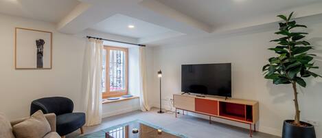 A modern living room with a sofa, a coffee table, a TV on a stand, a potted plant, and a window with curtains.