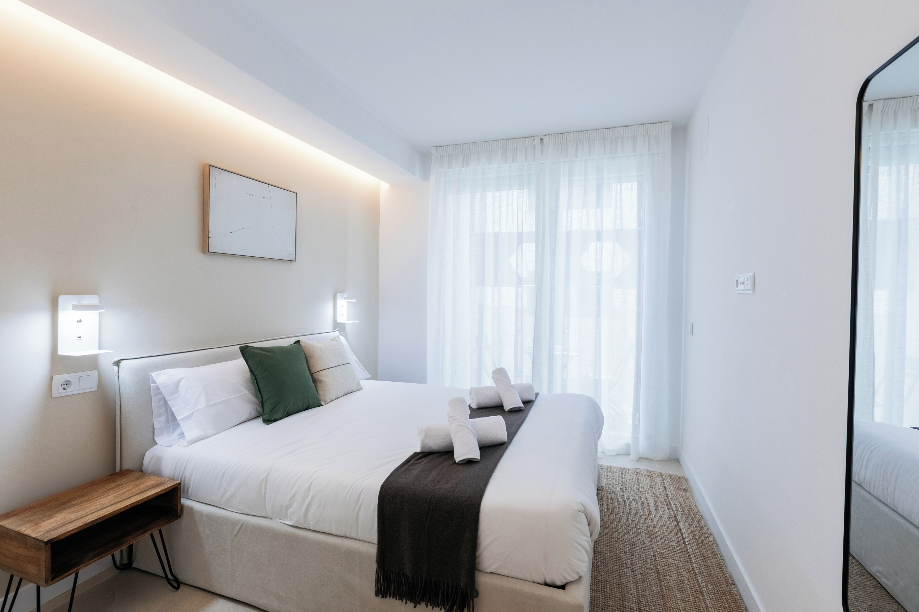 A hotel room with a bed, bedside table, wall-mounted light, and a window with sheer curtains.