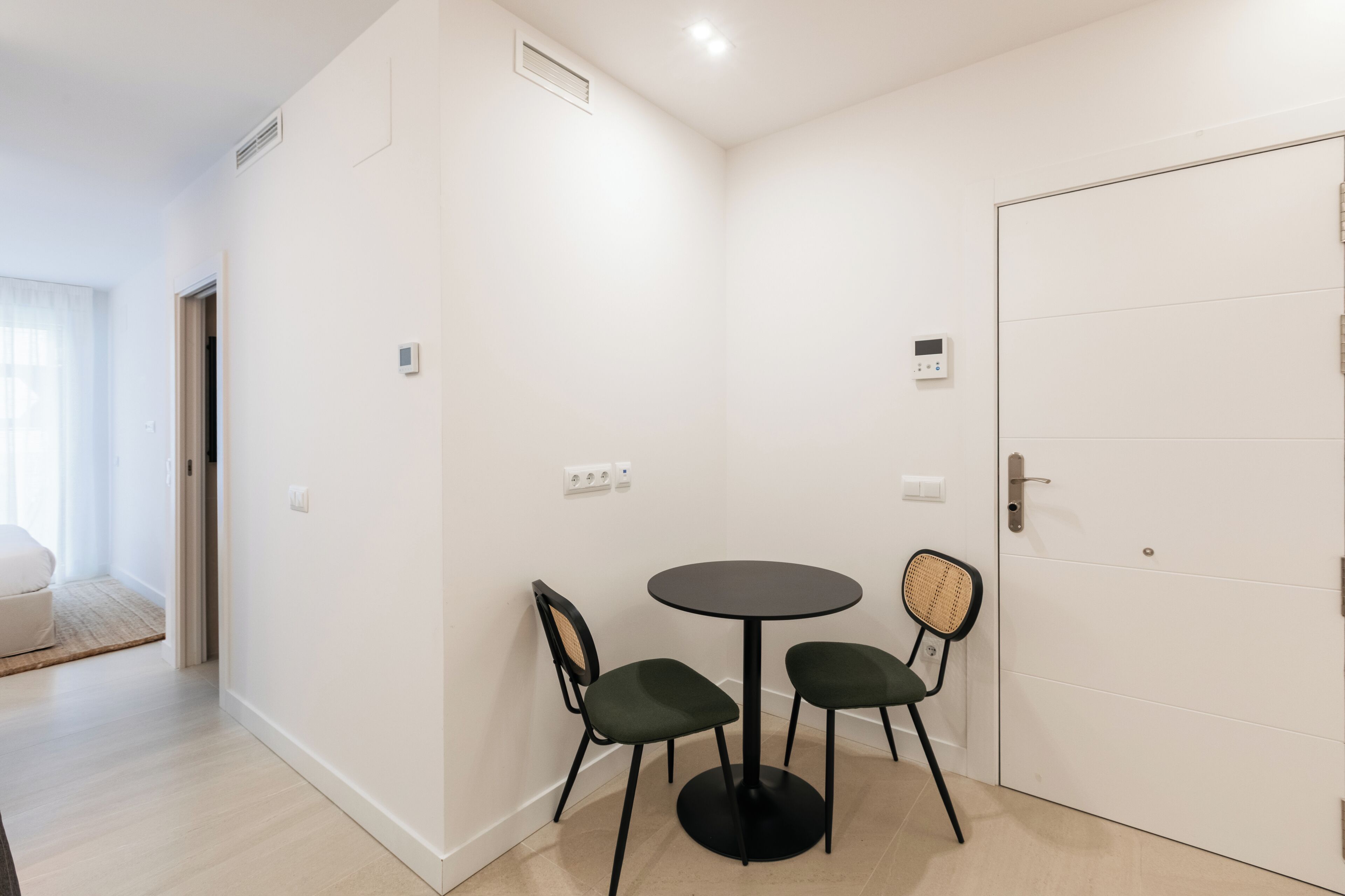 A room with a white door, a round table with two chairs, a small window with curtains, and a light switch on the wall.