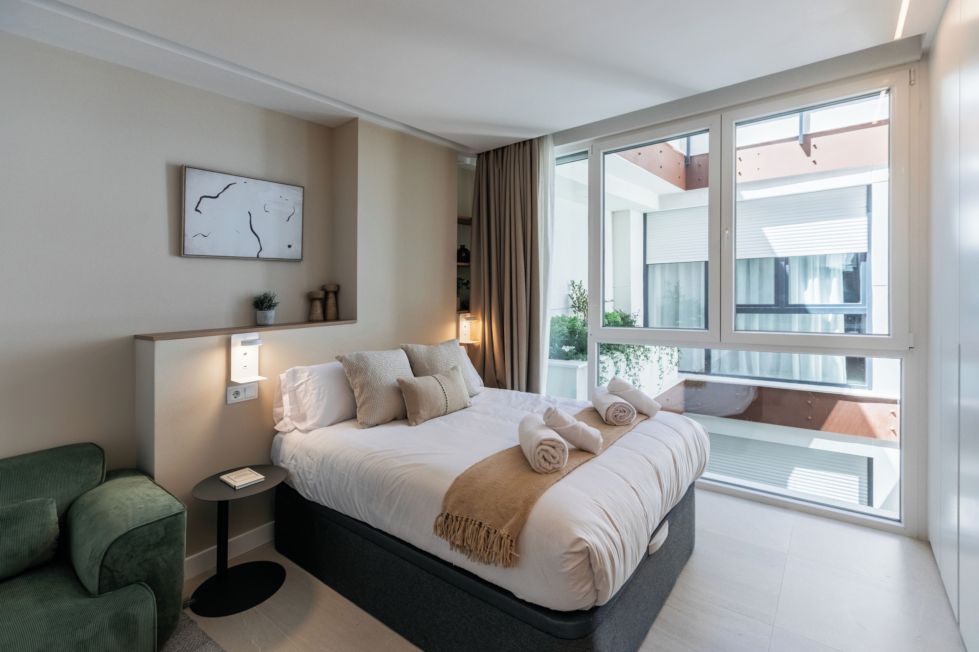 Una camera d'albergo moderna con un letto grande, un tavolino, una poltrona verde e un'ampia finestra con balcone.