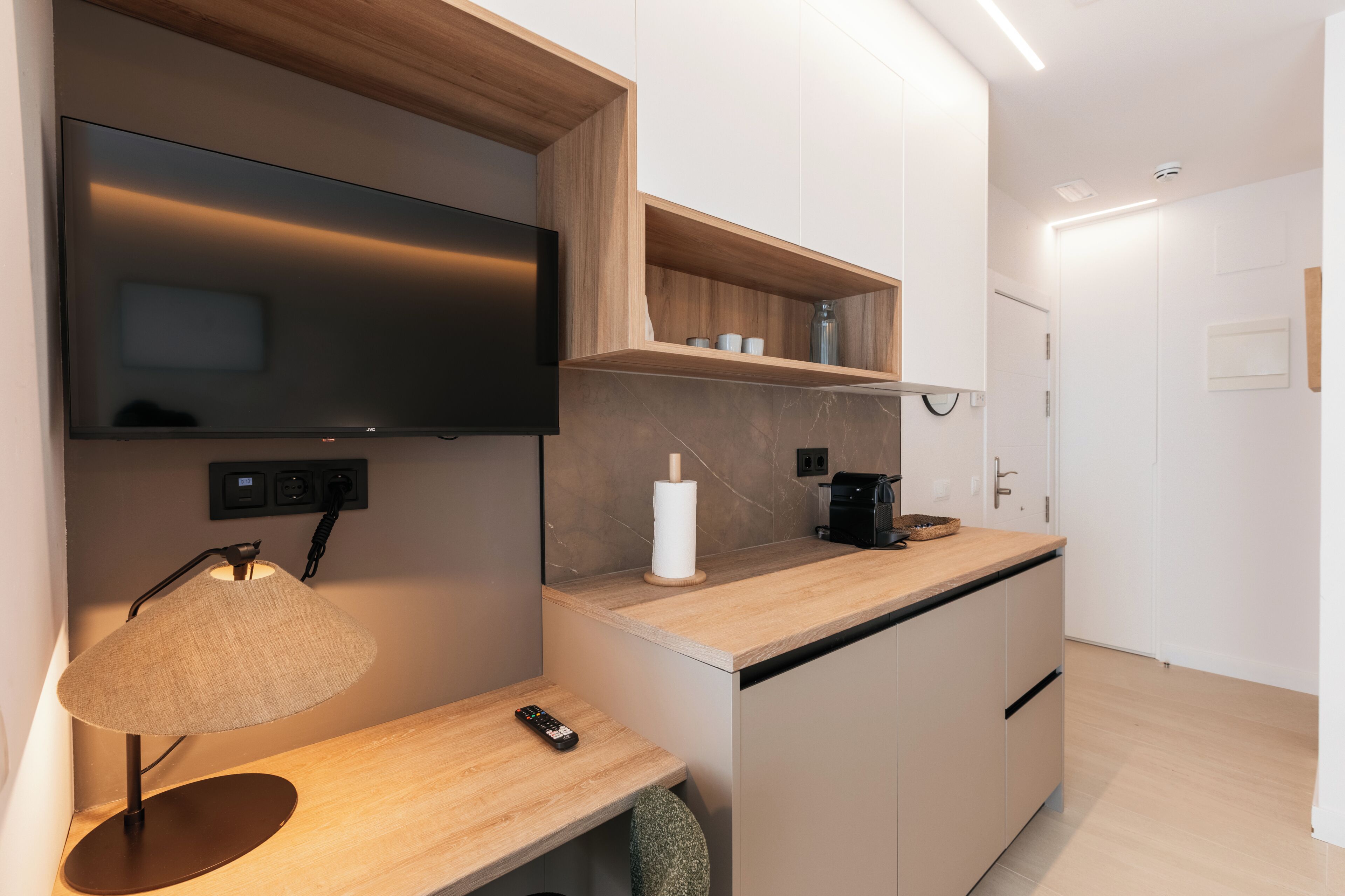A modern kitchen with a flat-screen TV, wooden shelves, a coffee maker, and a remote control on a wooden countertop.