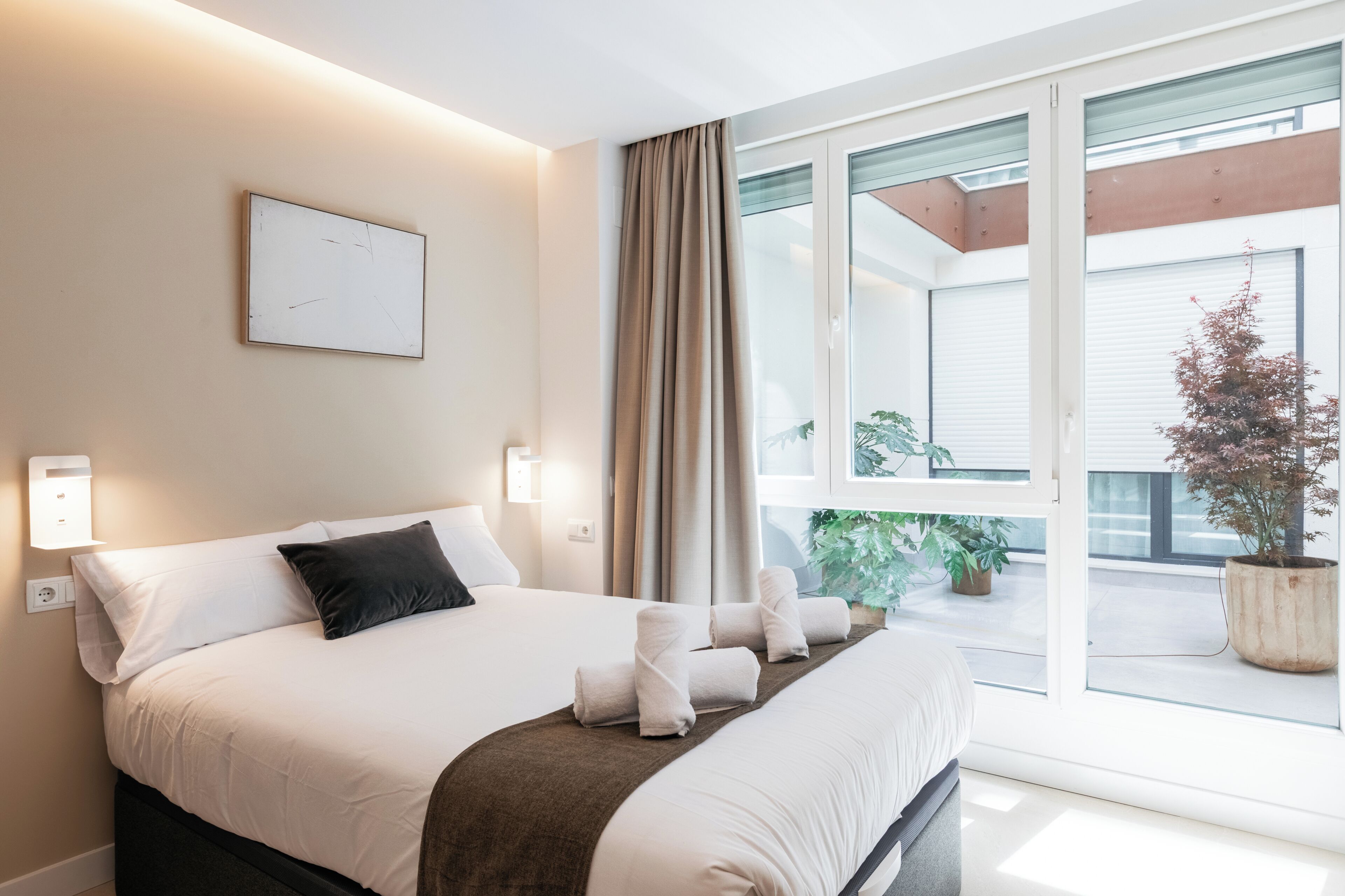Una habitación de hotel con cama, ventana con cortinas, planta en maceta y lámpara fijada a la pared.