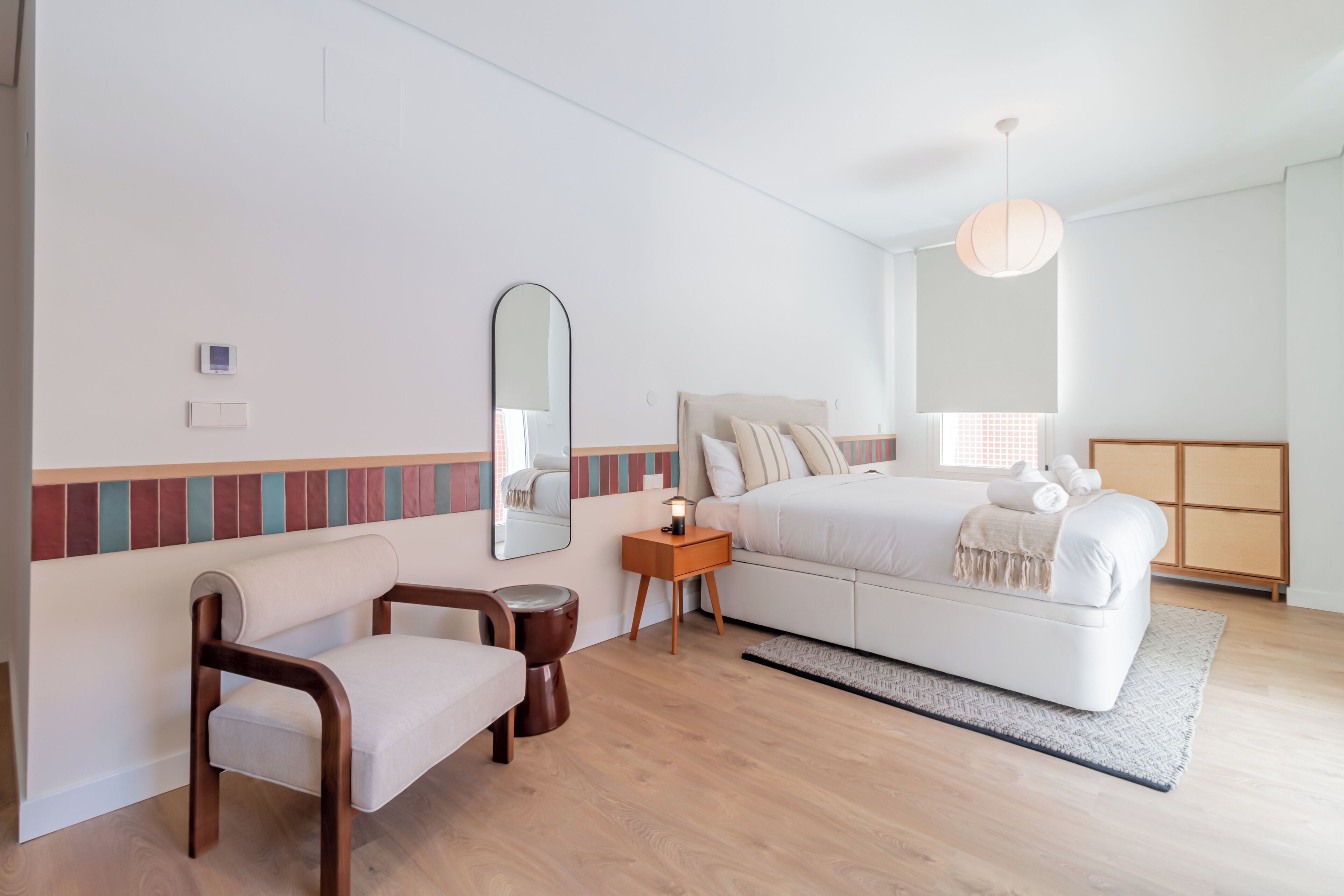 Un dormitorio con cama, mesita de noche, lámpara, silla y un taburete pequeño.