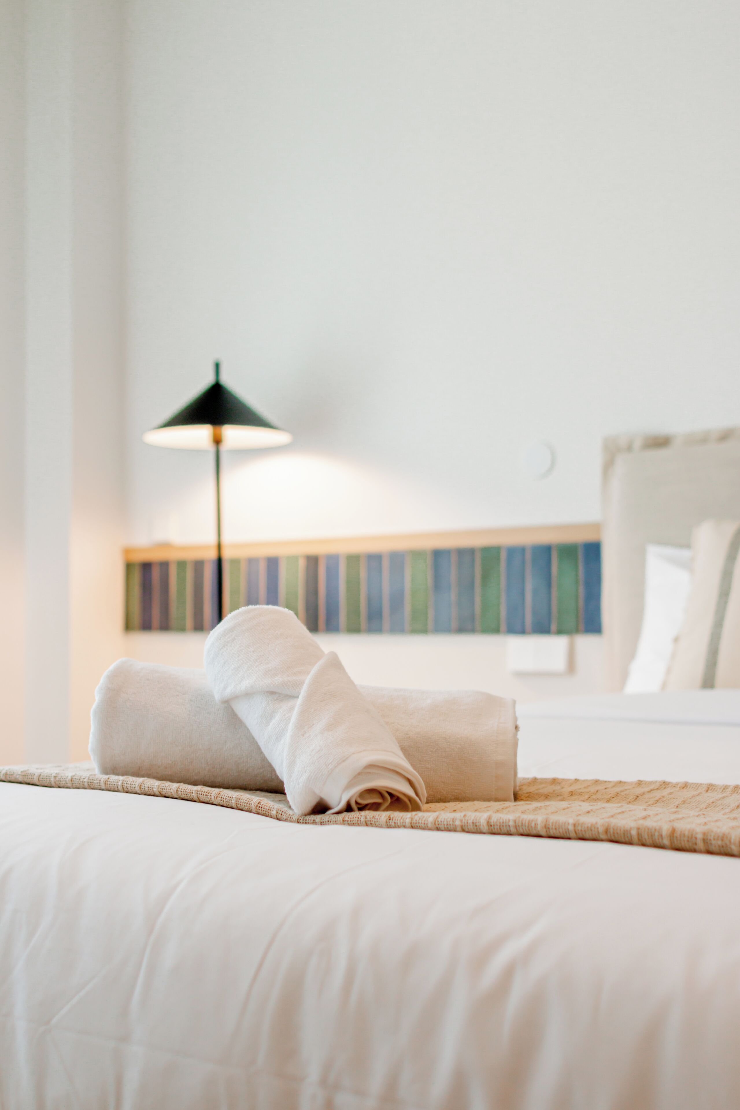 A neatly made bed with a rolled towel, a black floor lamp, and a striped wall decoration.