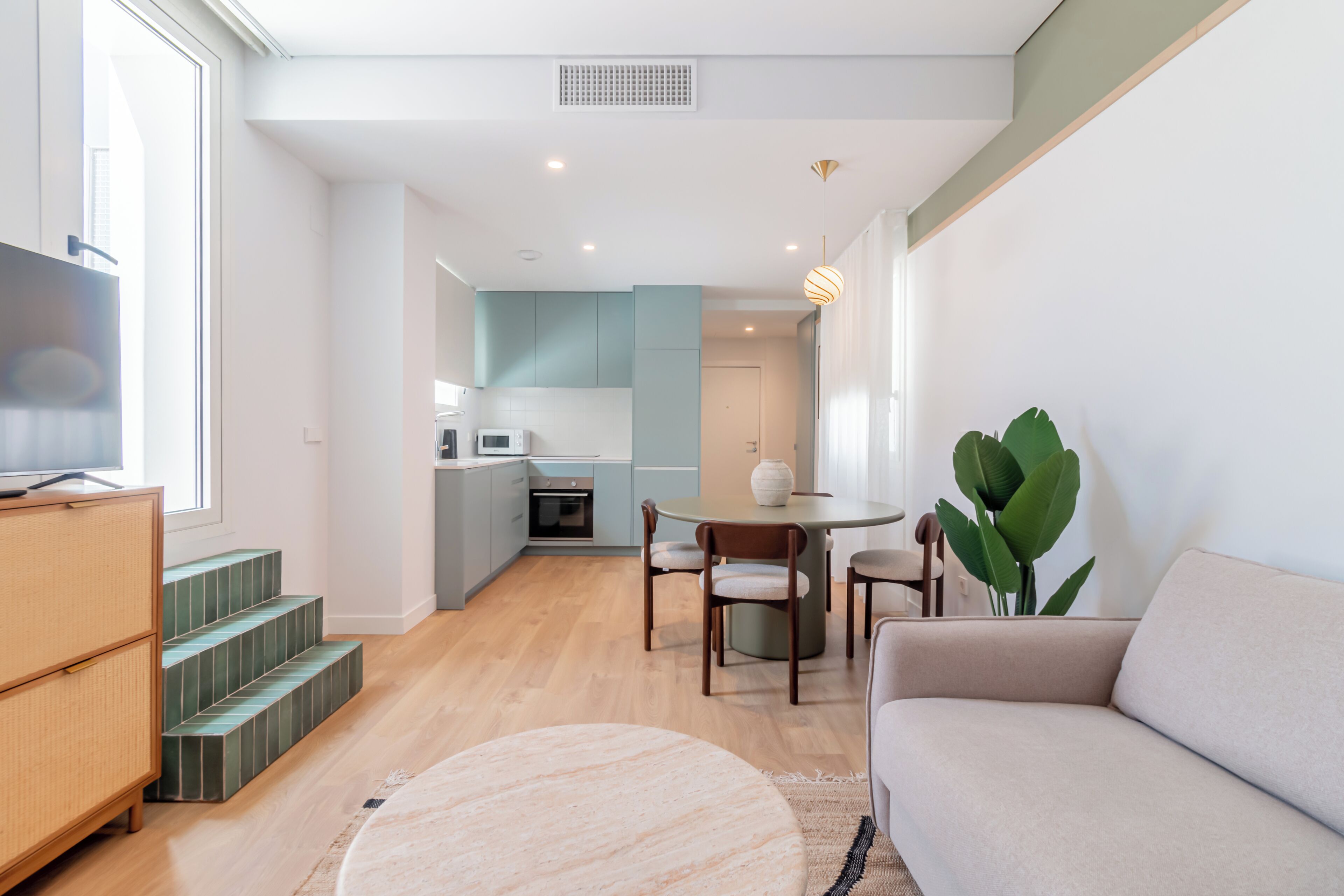 A modern living room with a sofa, a dining table with chairs, a kitchen area with cabinets, and a staircase.