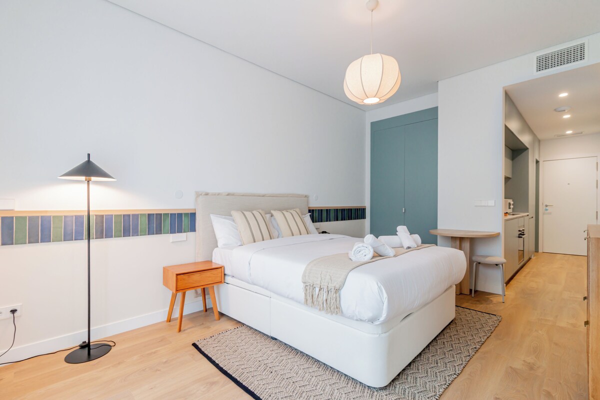Un dormitorio moderno con cama, mesita de noche, lámpara y un pequeño taburete.