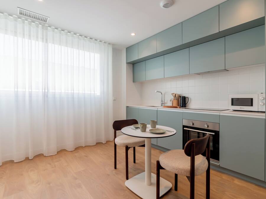 A modern kitchen with light blue cabinets, a round dining table with chairs, and a window with sheer curtains.
