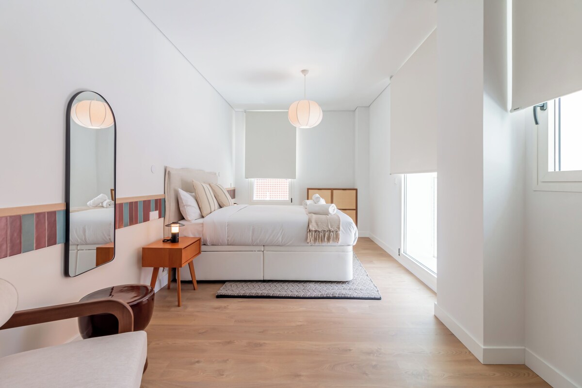 Un dormitorio moderno con cama, mesita de noche, espejo y vistas al exterior.