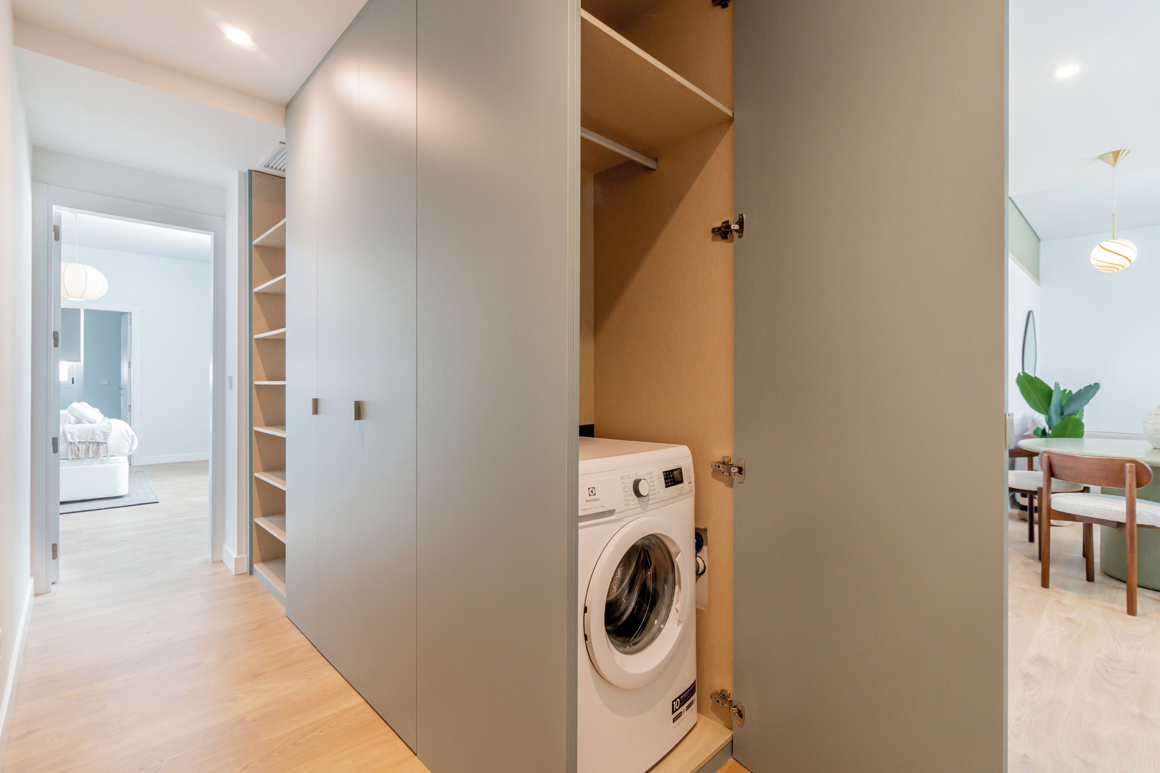 A modern laundry room with a built-in washer and dryer, a wooden floor, and a view into a dining area with a table and chairs.