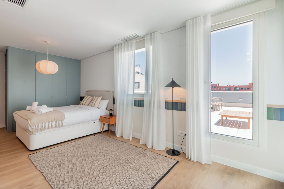Un dormitorio con cama, mesita de noche, lámpara y vistas al exterior.