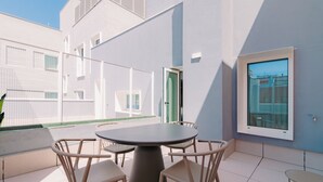 Et moderne udendørs spiseområde med et rundt bord og stole, udsigt til bygninger, og en dør.