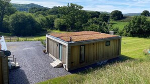 Een klein houten huisje met een groen dak, omgeven door een grindpad en gazons.