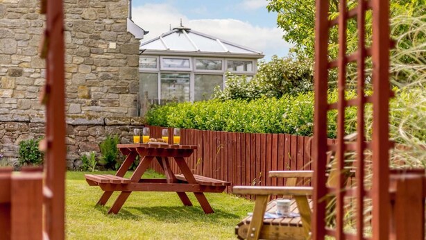 A patio with a wooden table and chairs, a stone wall, and a greenhouse in the background. - 2 Bed in Alnwick (oc-60464) (Boulmer)