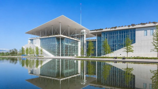 Un edificio moderno con fachada de vidrio, una masa de agua reflectante al frente y cielo despejado.