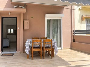 Un balcon avec une table et des chaises en bois, une porte-fenêtre coulissante et un petit coin cuisine visible à travers une porte ouverte.